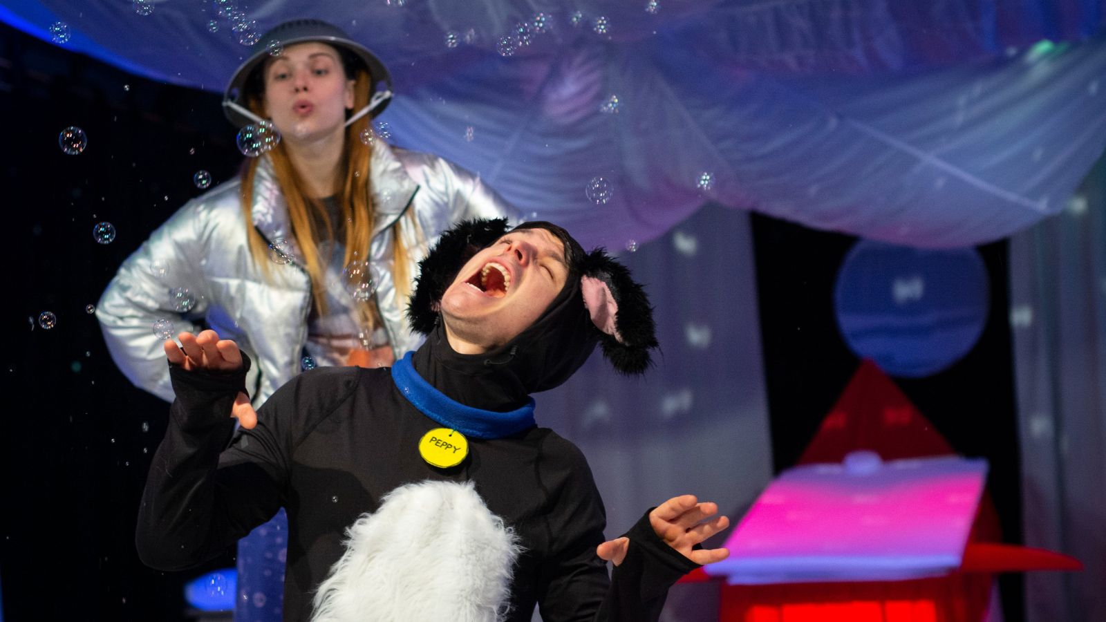 A fun performance where someone dressed as a dog is pretending to sing at the front of the stage, in front of someone else wearing a metal colander as a hat, as though they're dressing up like children, in a colourful environment